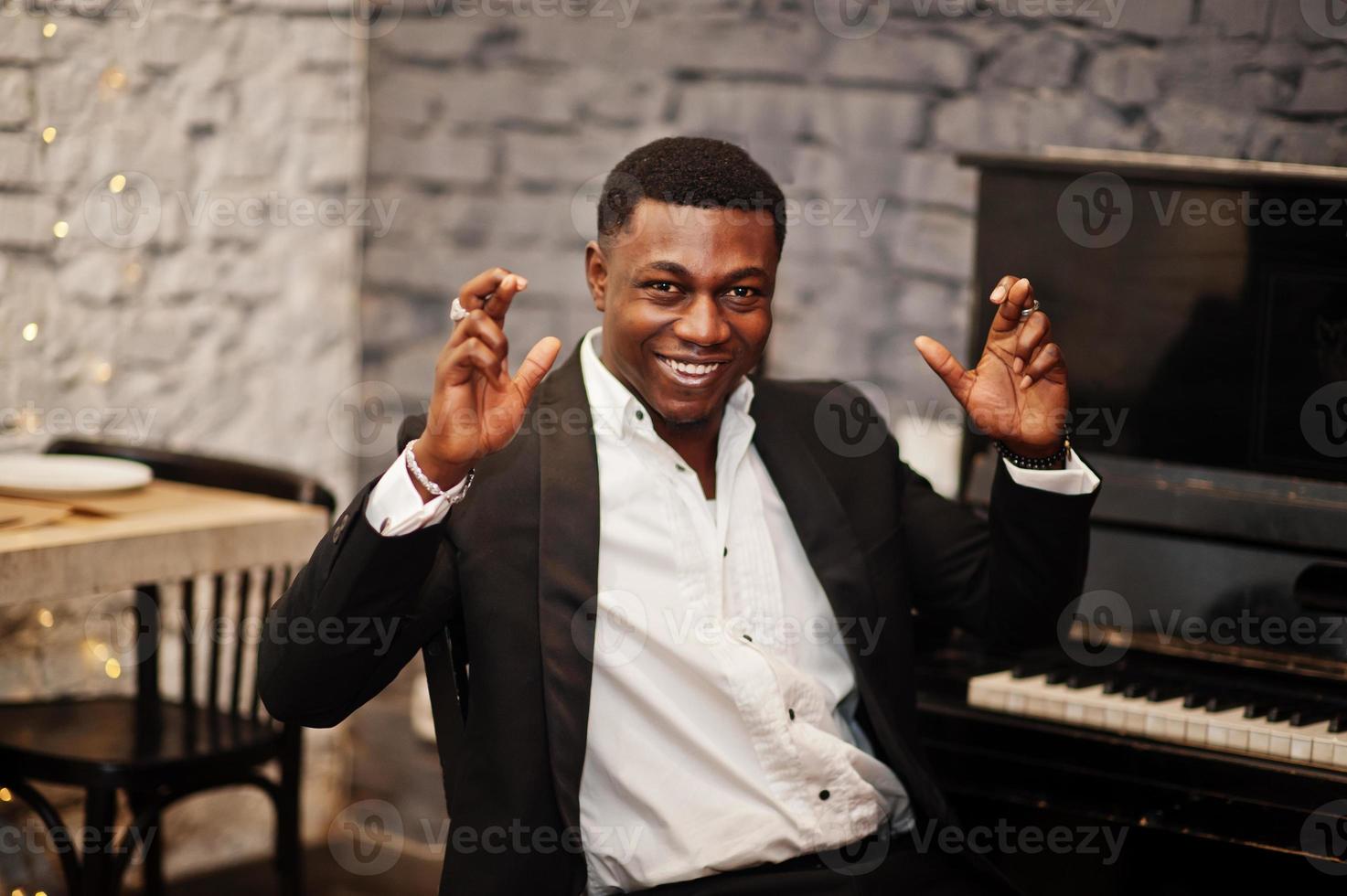 Strong powerful african american man in black suit sitting against piano and cross his fingers for luck. photo