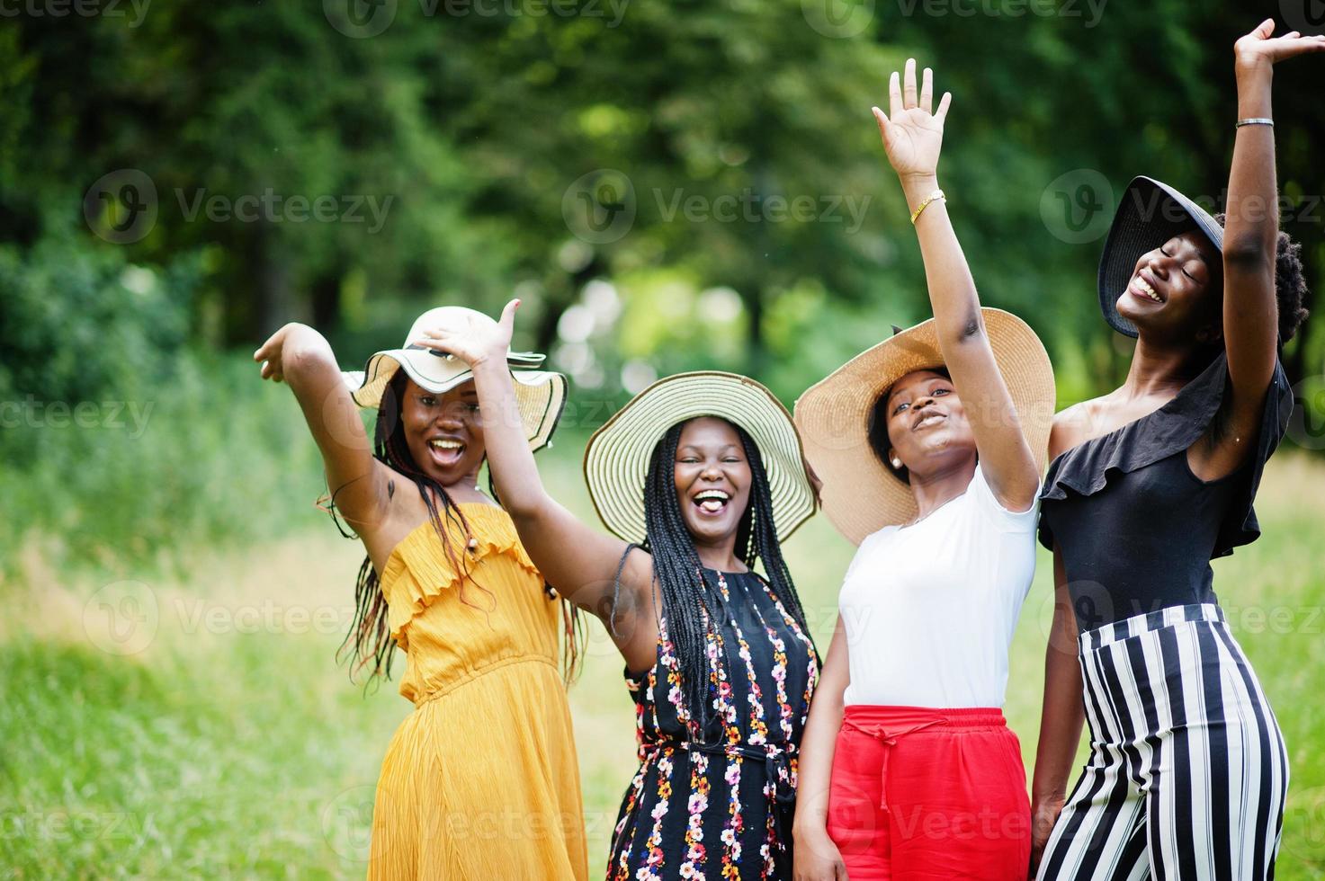 Group of four gorgeous african american womans wear summer hat spending time at green grass in park. photo