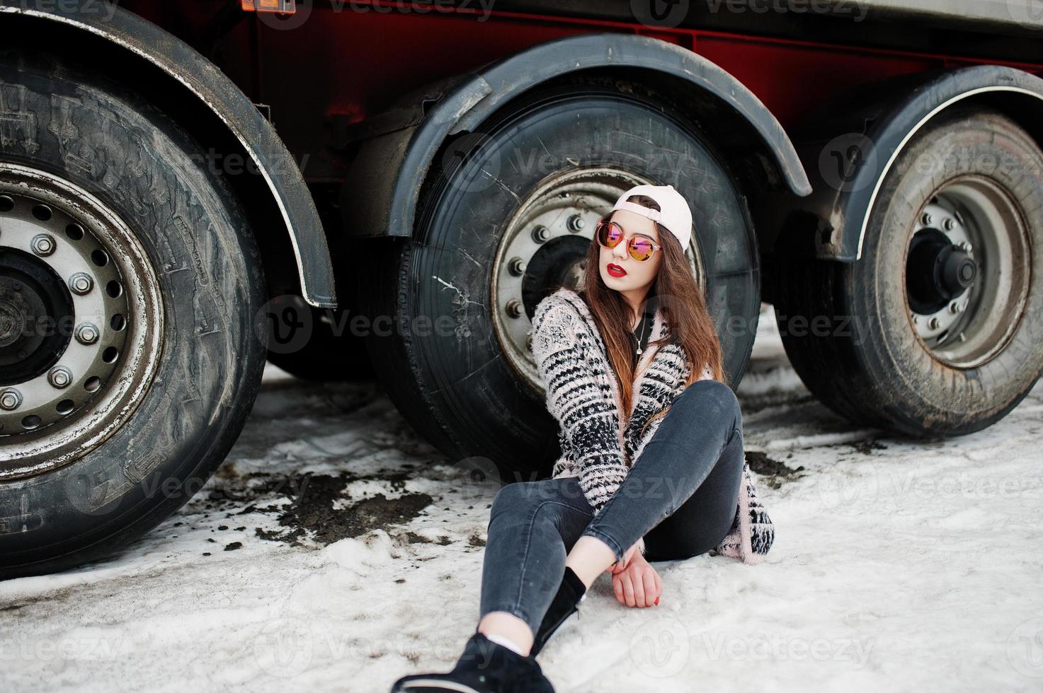 chica casual elegante morena con gorra y gafas de sol sentada contra las ruedas del camión. foto