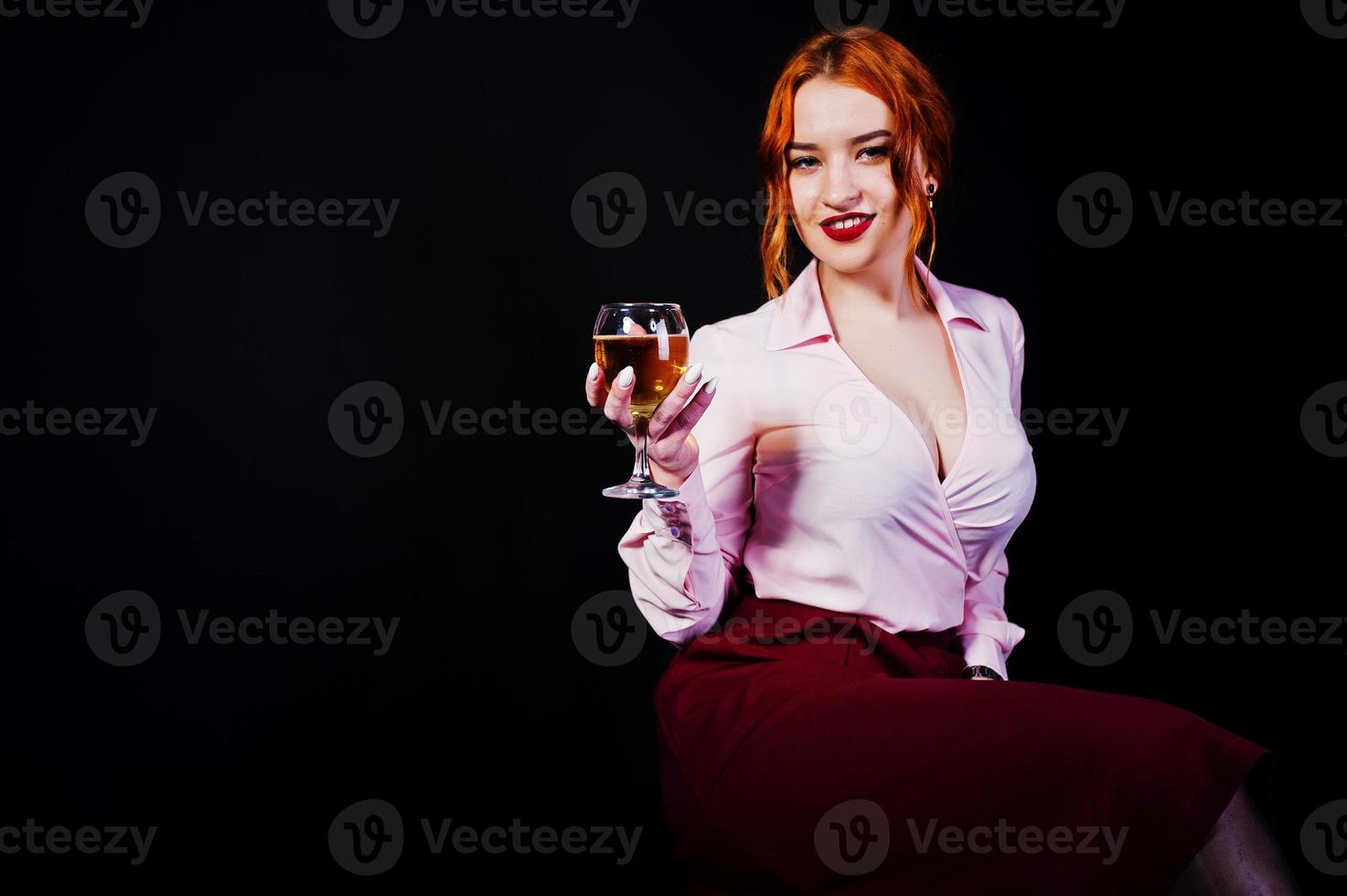 Hermosa chica pelirroja con blusa rosa y falda roja con una copa de vino a mano aislada en negro. foto