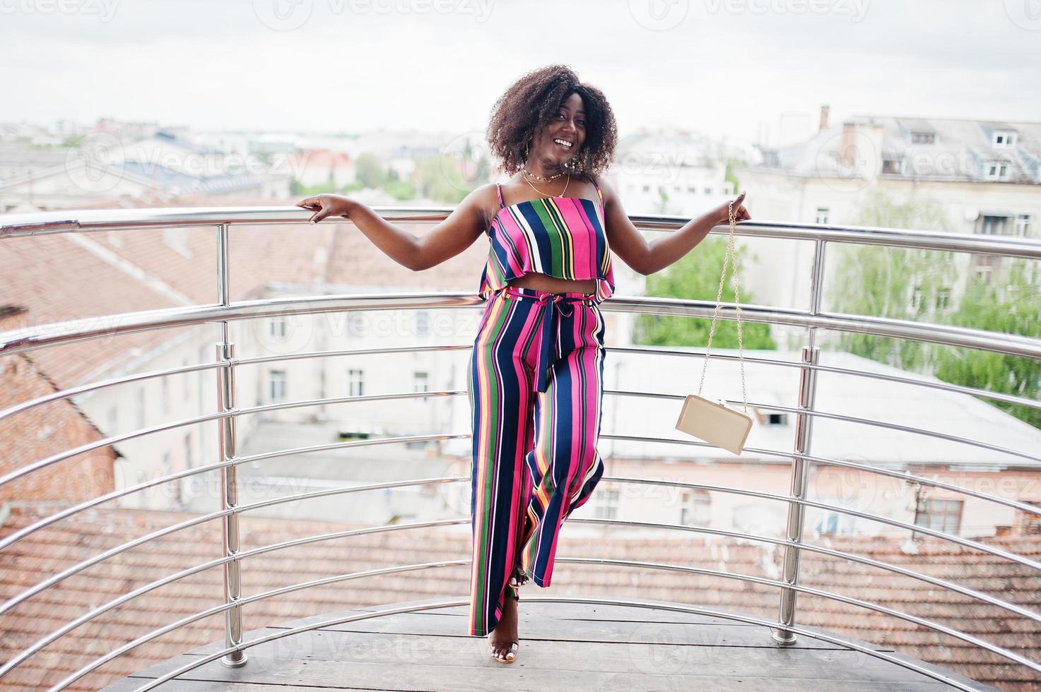 Fashionable african american woman in pink striped jumpsuit, with handbag posed in the french balcony. photo