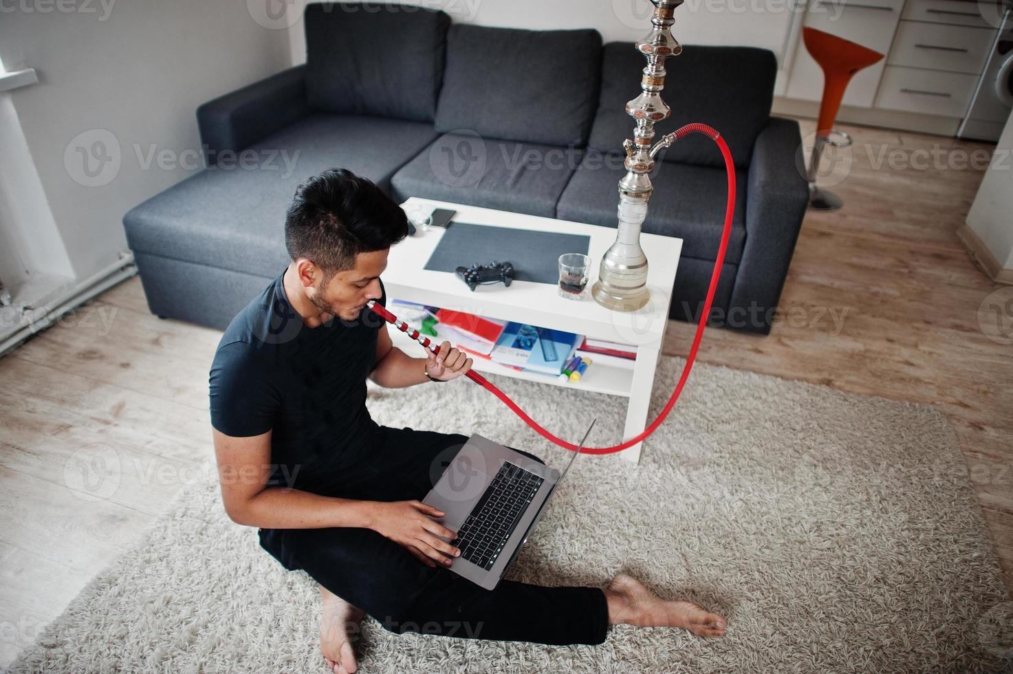 Handsome and fashionable indian man in black sitting at room, smoking hookah and working on laptop. photo