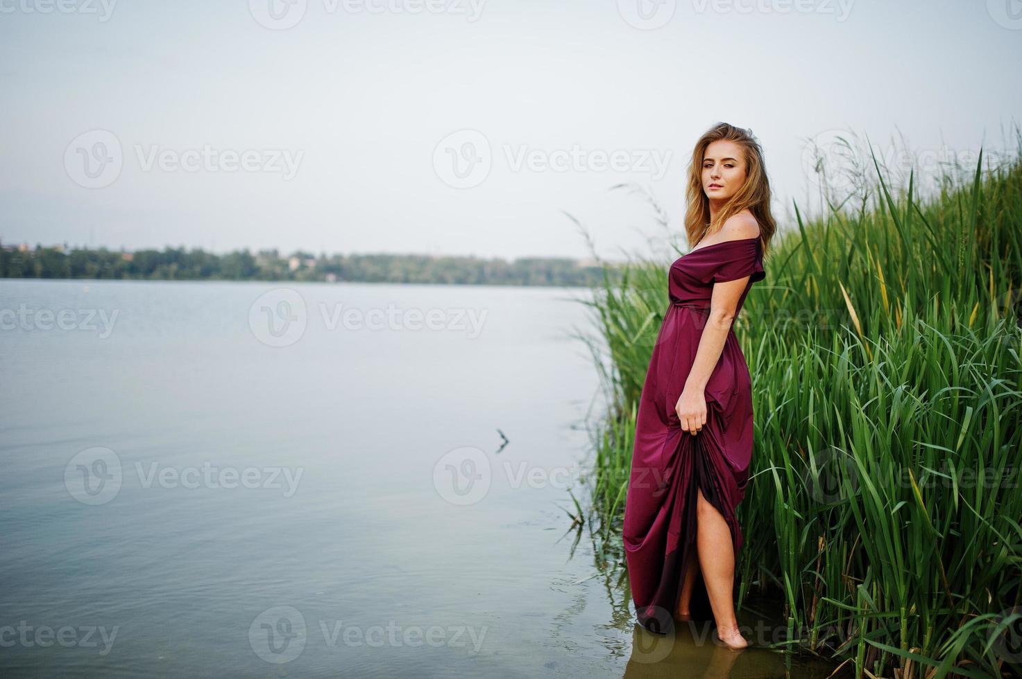 Blonde sensual woman in red marsala dress standing in water of lake with reeds. photo