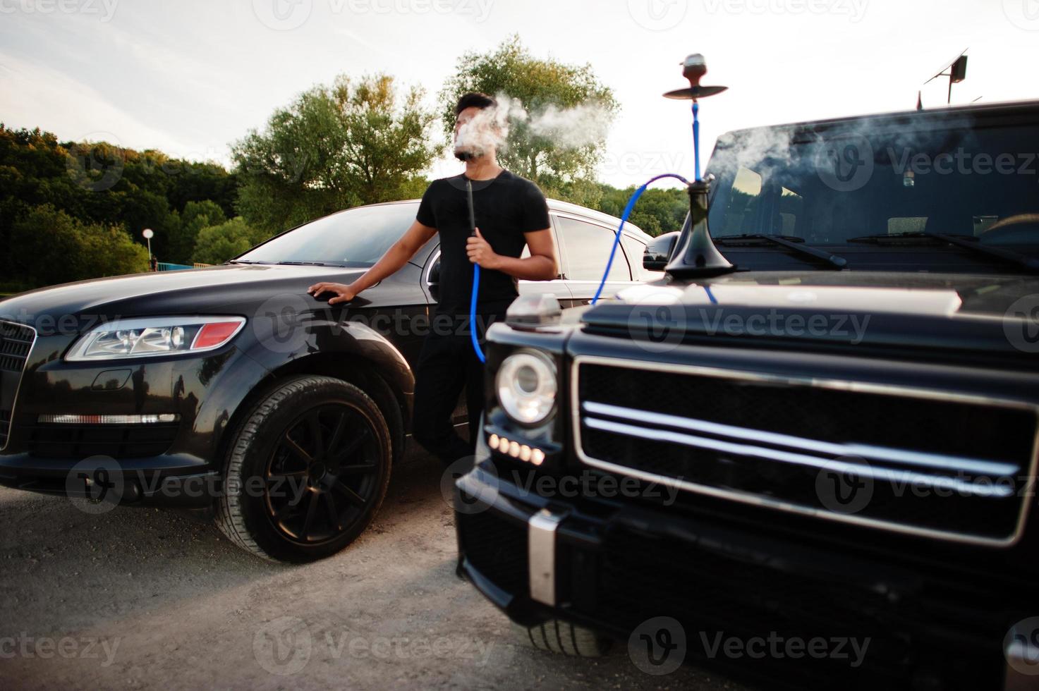 Asian man wear on all black posed near suv car and smoke hookah. photo