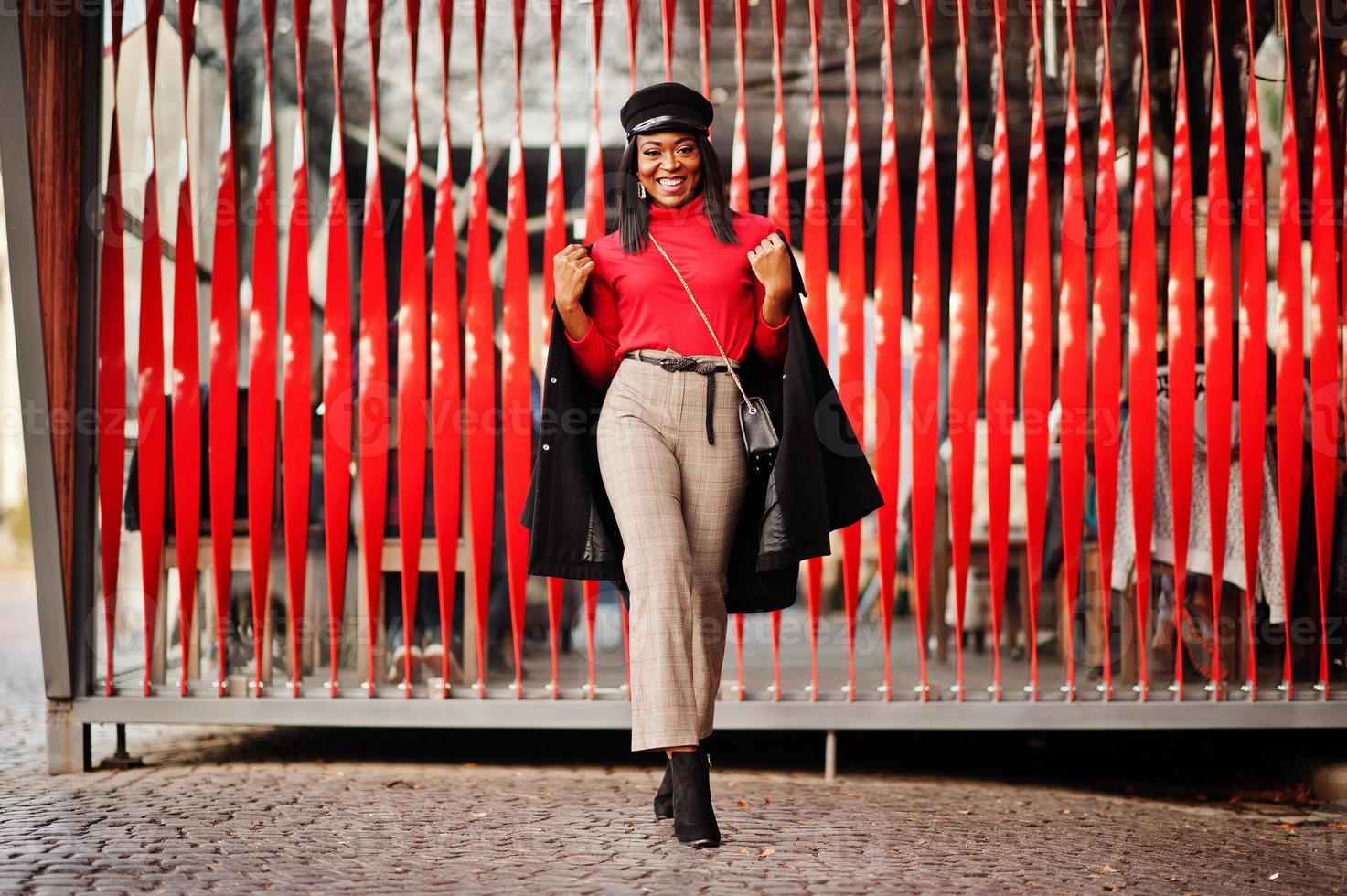 African american fashion girl in coat and newsboy cap posed at street against red background. photo