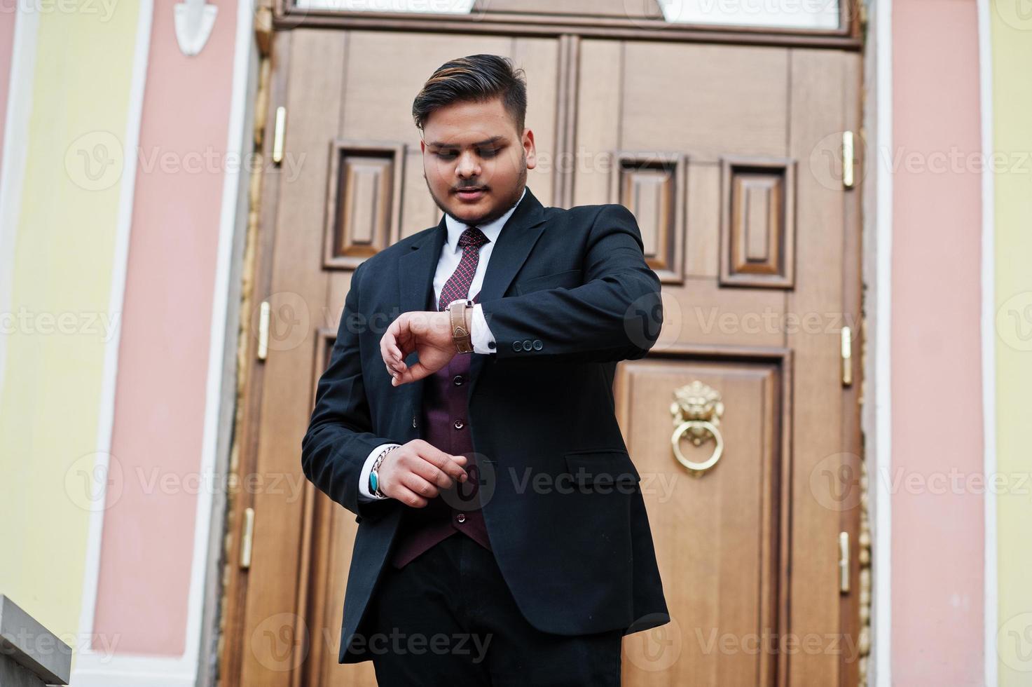 hombre de negocios indio con estilo en ropa formal de pie contra la puerta en el centro de negocios y mira sus relojes en la mano. foto