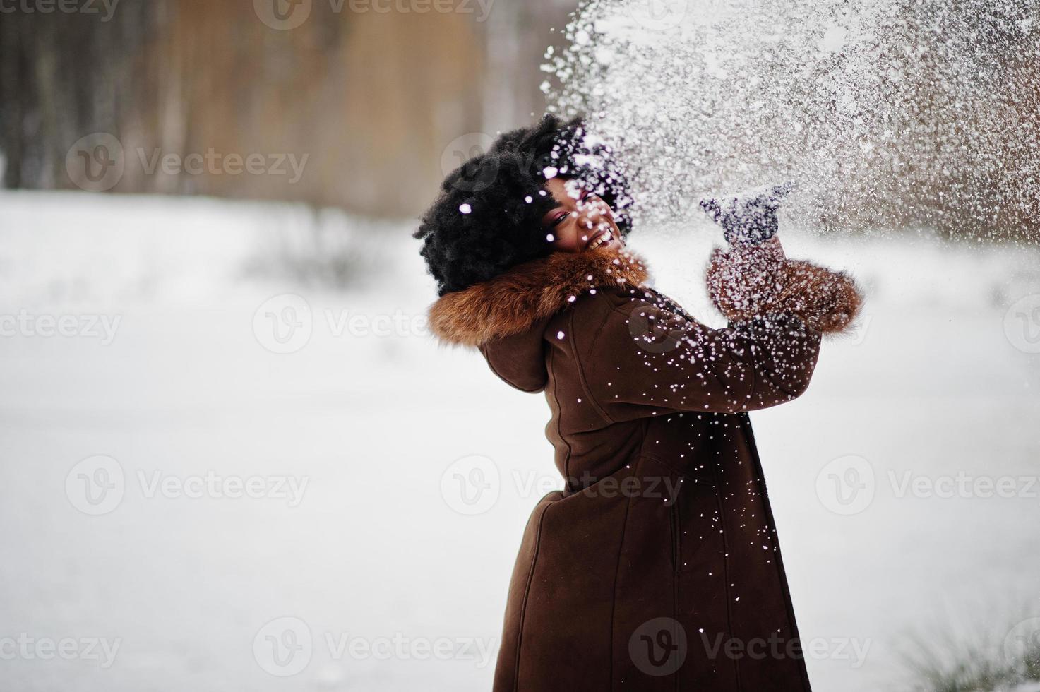 Curly hair african american woman wear on sheepskin coat and gloves posed at winter day throws up snow. photo