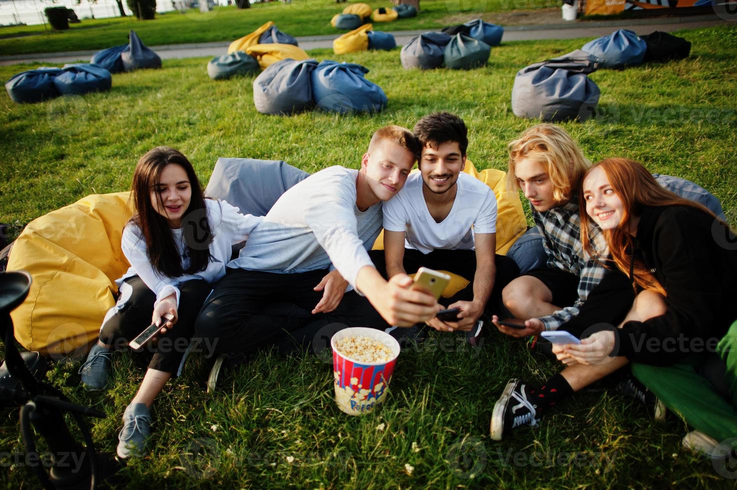 Young multi ethnic group of people watching movie at poof in open air cinema and making selfie on phone. photo