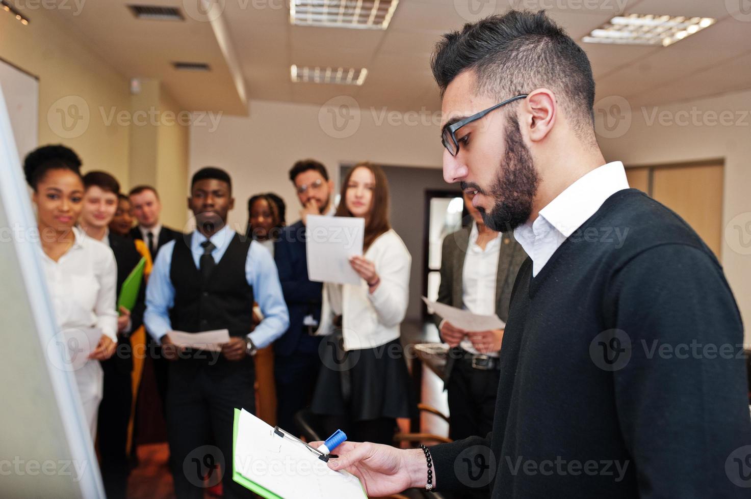 Arabian business coach presenting report standing near whiteboard pointing on sales statistic shown on diagram and chart teach diverse multiracial company members gathered together in conference room. photo