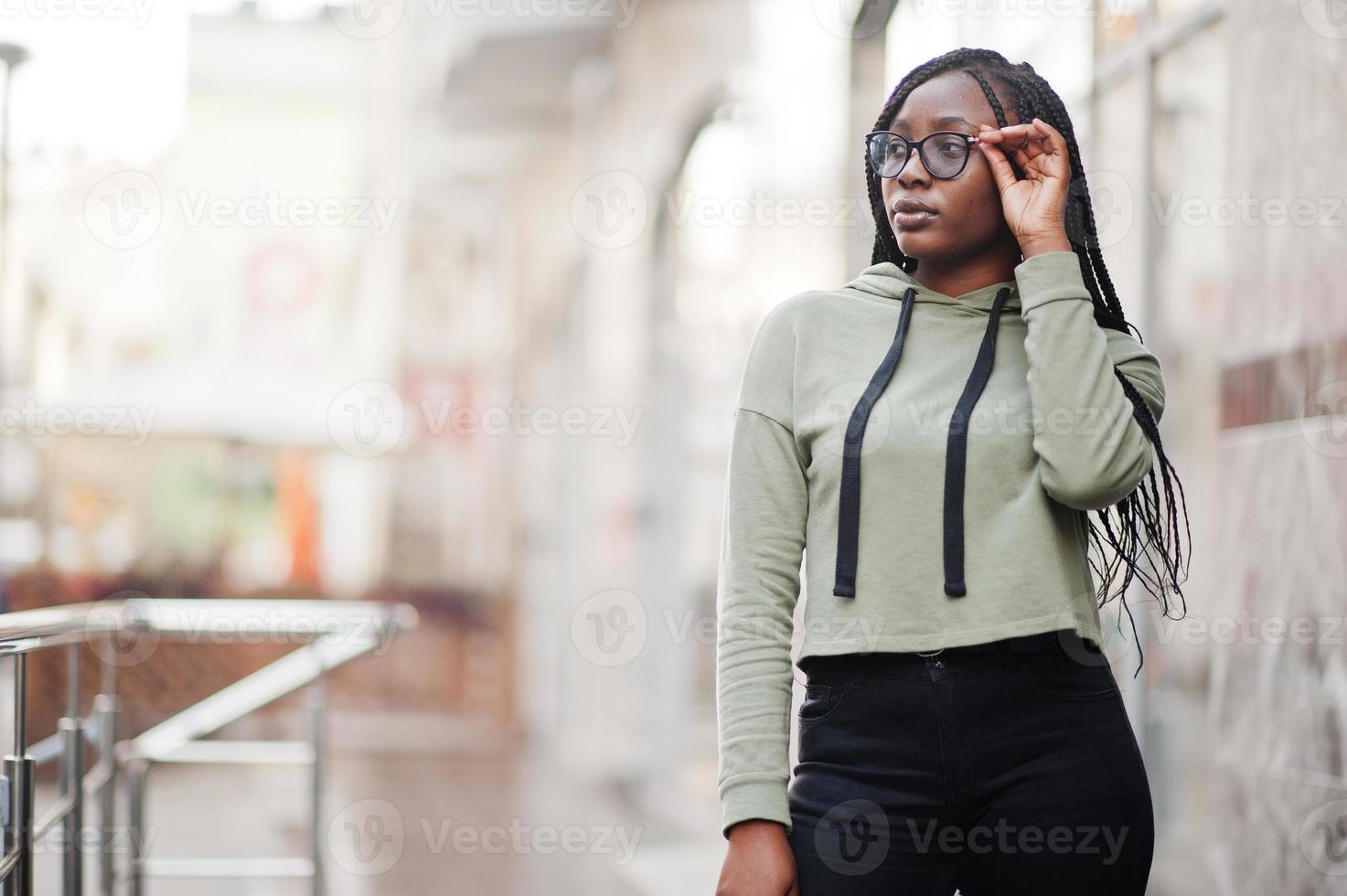 City portrait of positive young dark skinned female wearing green hoody and eyeglasses. photo