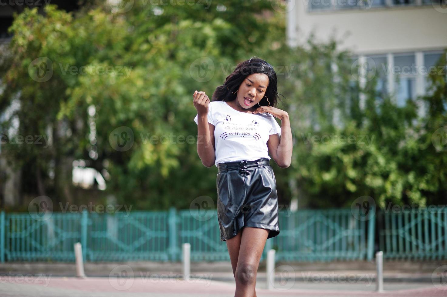 Modelo delgado afroamericano de piel oscura posado en pantalones cortos de cuero negro y camiseta blanca. foto