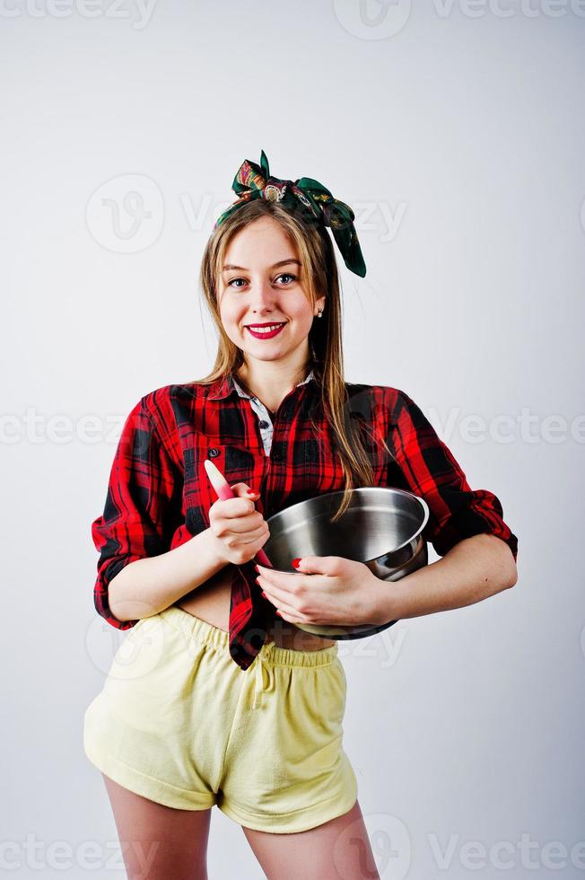 joven ama de casa divertida en camisa a cuadros y pantalones cortos amarillos pin up estilo con cacerola y cuchara de cocina aislado sobre fondo blanco. foto