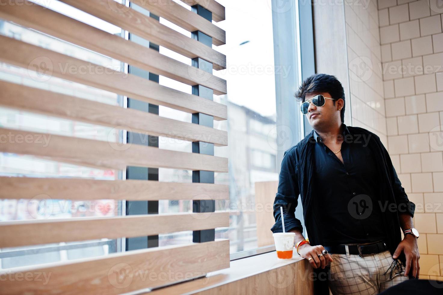 un hombre indio elegante con gafas de sol se queda cerca de la ventana con una taza de café en un café de comida rápida. foto
