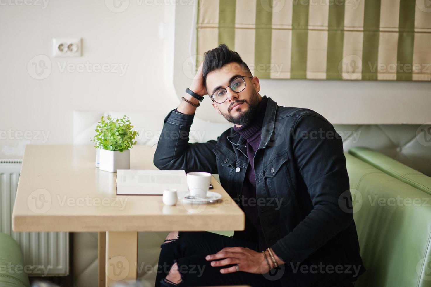 el hombre árabe usa una chaqueta de jeans negros y anteojos sentados en un café, lee un libro y bebe café. chico modelo árabe elegante y de moda. foto