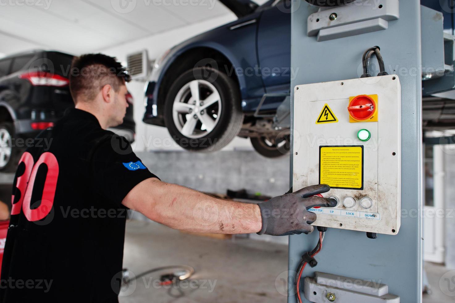 Car repair and maintenance theme. Mechanic in uniform working in auto service, press button to lifting auto. photo