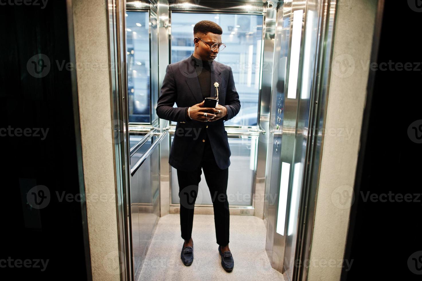 hombre afroamericano de moda con traje y gafas con teléfono móvil y una taza de café en las manos posado dentro del ascensor. foto