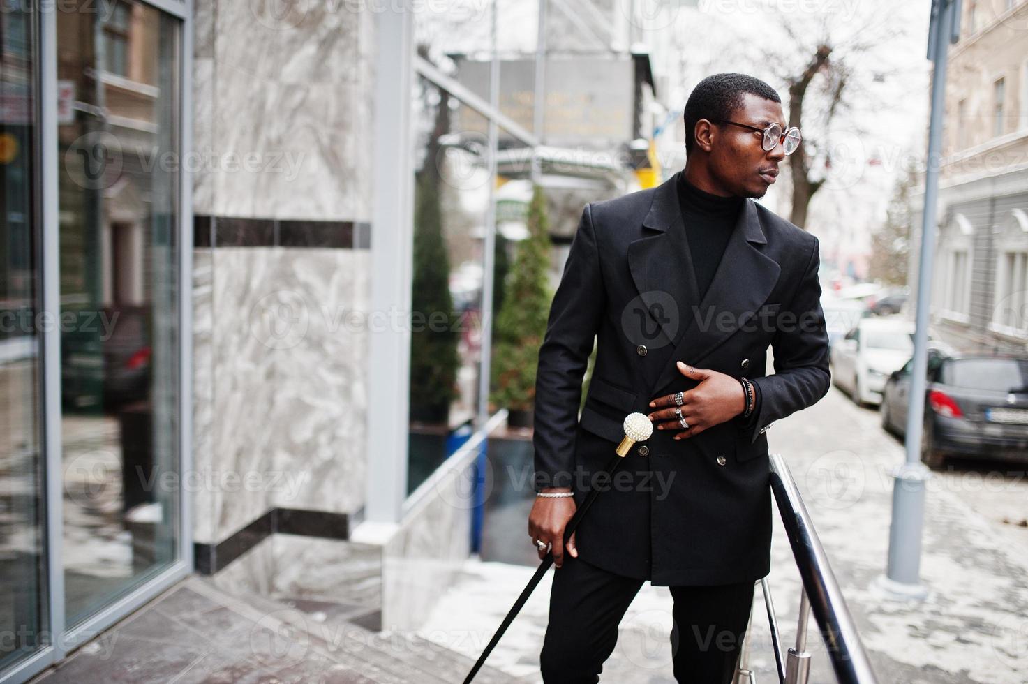 elegante caballero afroamericano con elegante chaqueta negra, sosteniendo un bastón retro como matraz de caña o bastón basculante con mango de bola de diamante dorado. rico hombre afro de moda. foto