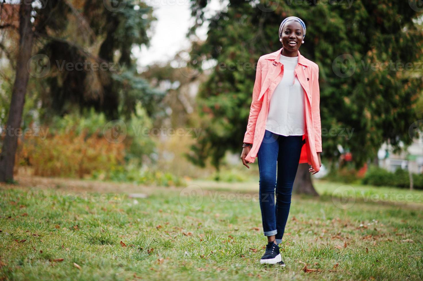 Young modern fashionable, attractive, tall and slim african muslim woman in hijab or turban head scarf and pink coat posed at park. photo