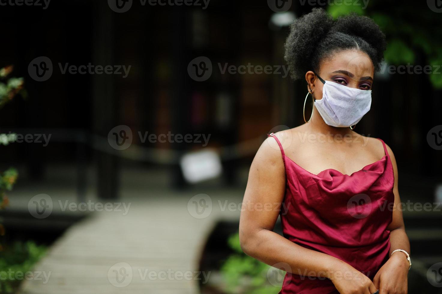 Covid-19, infectious virus. African woman with curly hair, wears red silk dress and medical disposable mask, cares about her health and protects in dangerious situation. photo