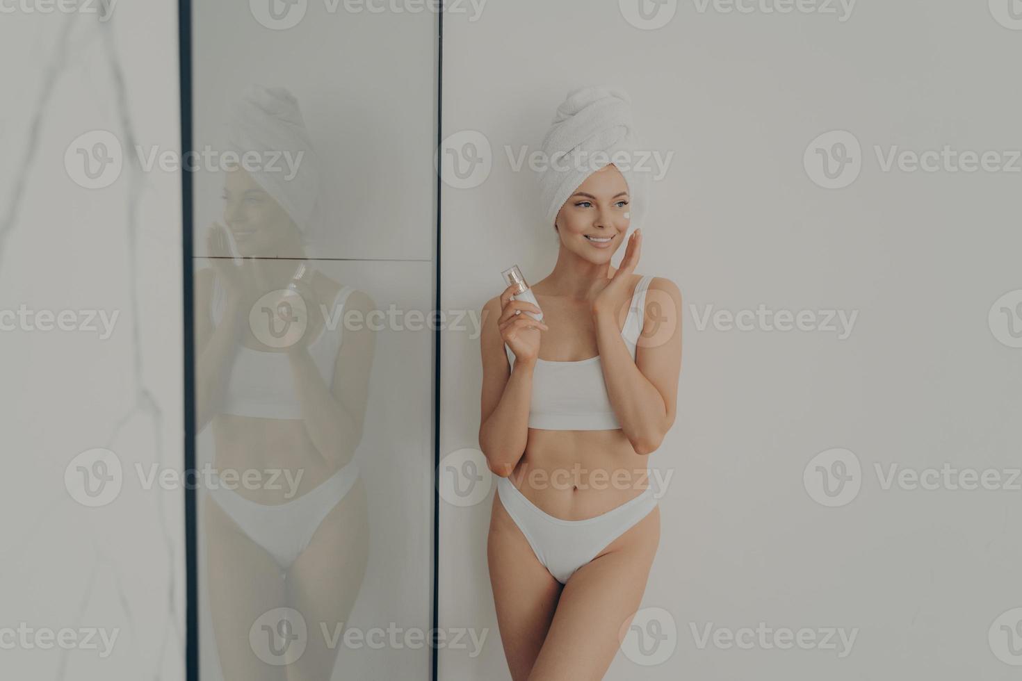 Beautiful woman standing in bathroom after morning shower and applying moisturizing cream on face photo