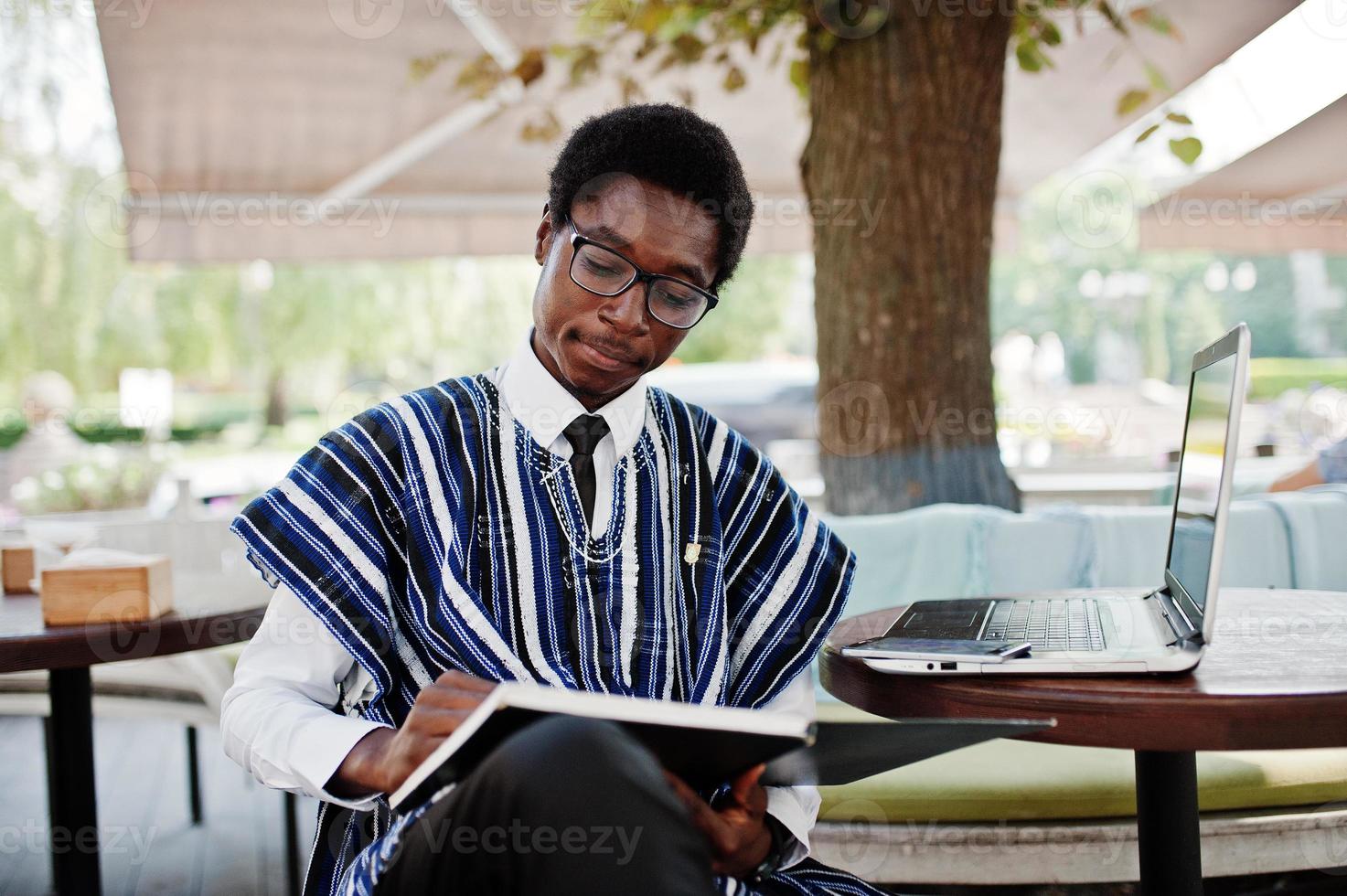 African man in traditional clothes and glasses sitting behind laptop at outdoor caffe and looking on his notebook. photo