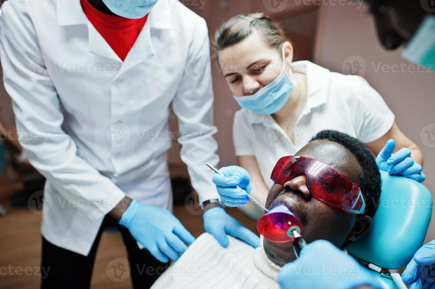 equipo multirracial de médicos dentistas. paciente afroamericano con gafas protectoras uv. sus dientes tratados con la ayuda de una lámpara de luz de curado ultravioleta dental y un espejo dental. foto