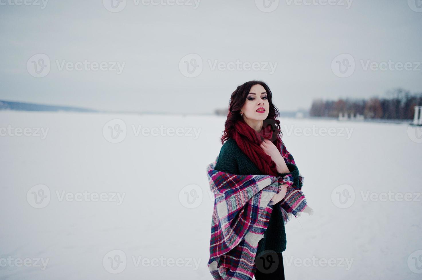 Brunette girl in green sweater and red scarf with plaid outdoor frozen lake on evening winter day. photo