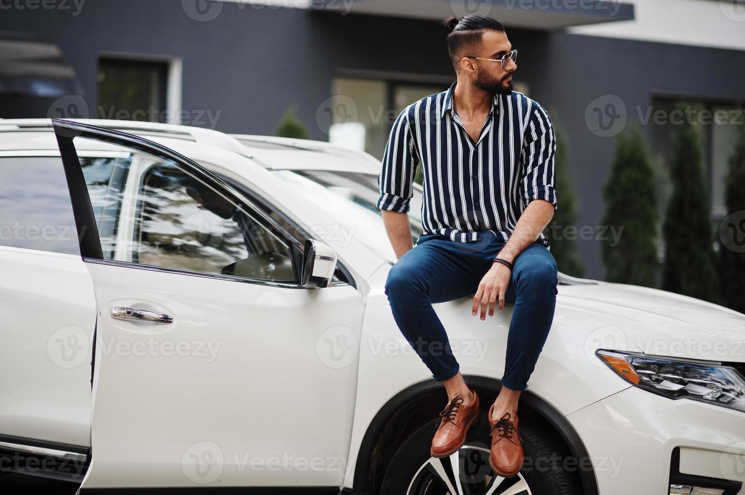 Successful arab man wear in striped shirt and sunglasses sitting on the hood of white suv car. Stylish arabian men in transport. photo