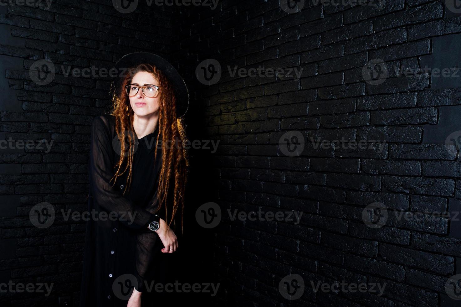 sesión de estudio de una chica de negro con rastas, anteojos y sombrero sobre fondo de ladrillo. foto