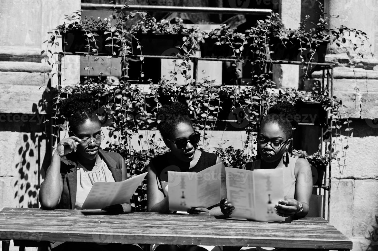 Three stylish african american womans posed at sunny summer day outdoor, sitting on table of restaurant with menu at hands. photo