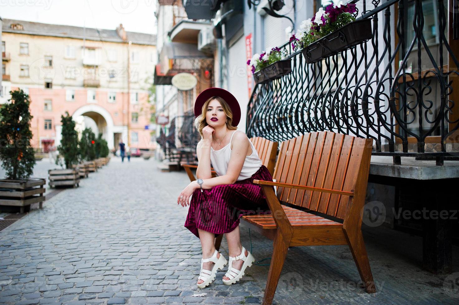 chica modelo rubia de moda y hermosa con elegante falda de terciopelo rojo, blusa blanca y sombrero, posada en un banco en la calle de la ciudad. foto