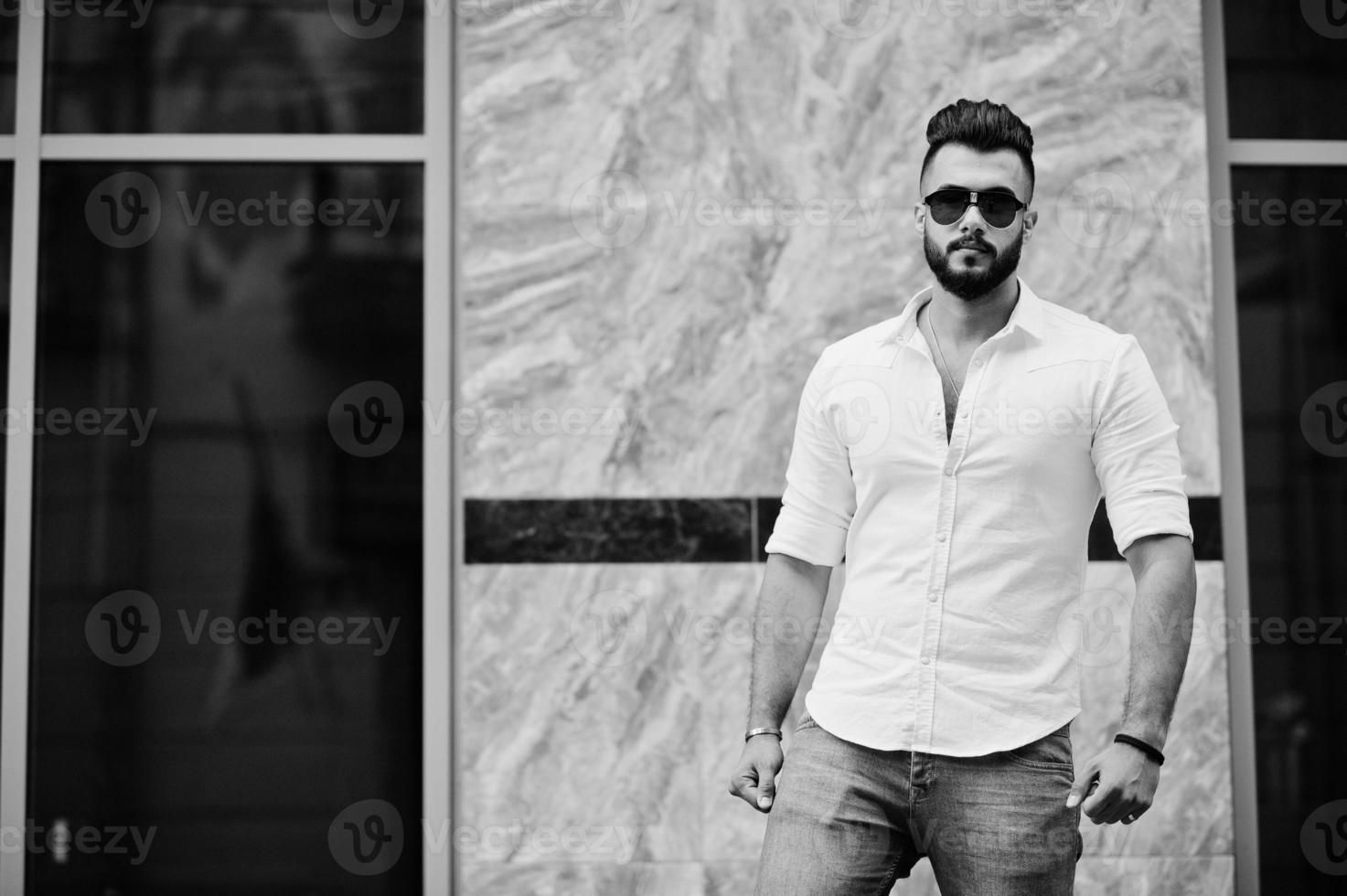 Stylish tall arabian man model in white shirt, jeans and sunglasses posed at street of city. Beard attractive arab guy. photo