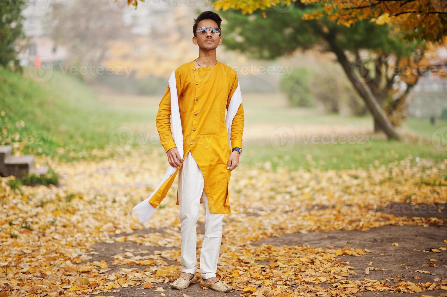 Indian stylish man in yellow traditional clothes with white scarf, sunglasses posed outdoor against autumn leaves tree. photo