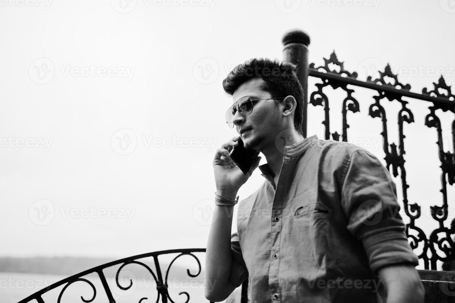 Indian man at red shirt and sunglasses posed outdoor and speaking on phone. Black and white photo. photo