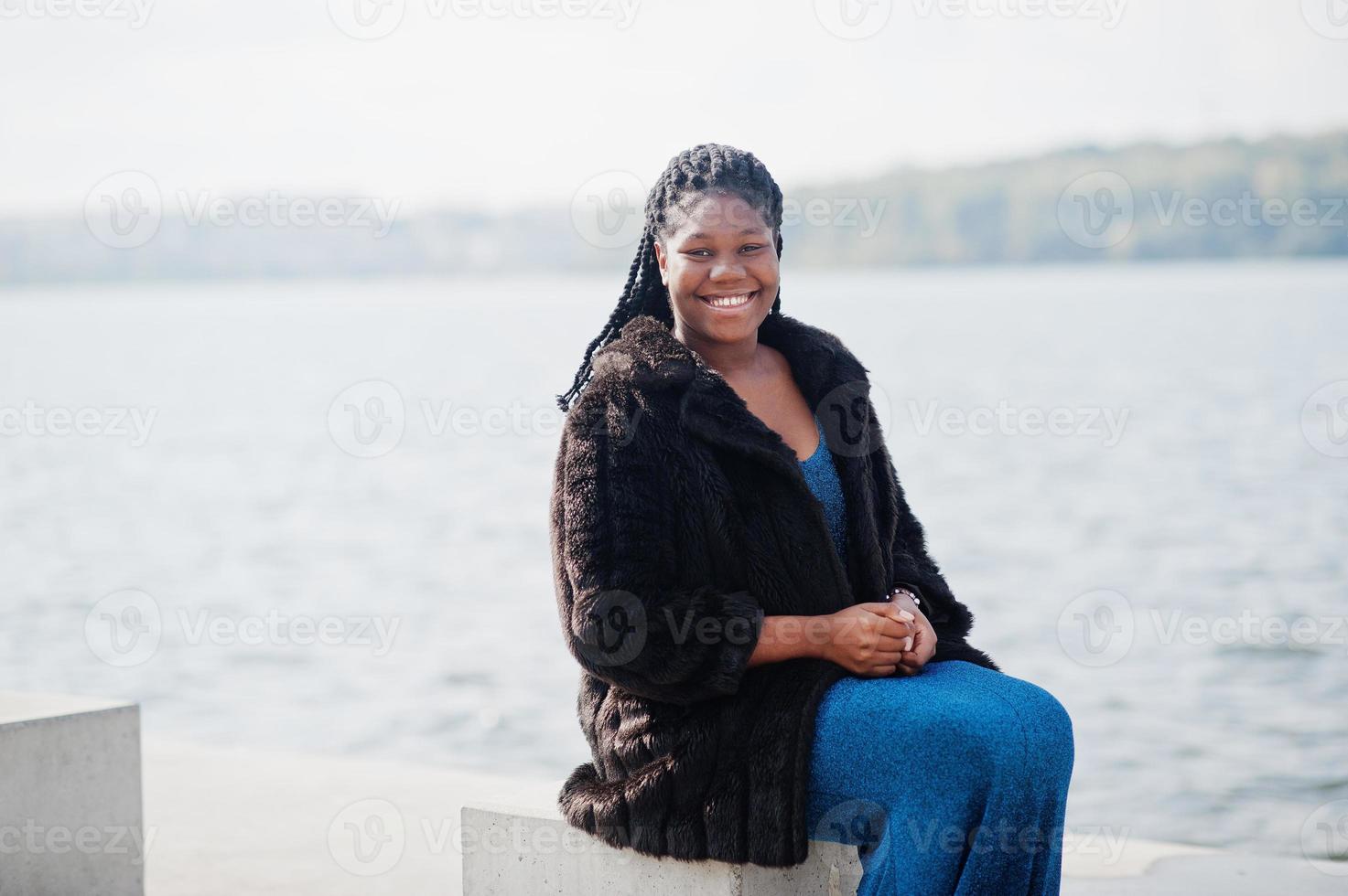 modelo afroamericano de piel oscura de talla grande posado con un vestido azul brillante y un abrigo de piel negro sentado en un cubo de piedra frente al mar. foto