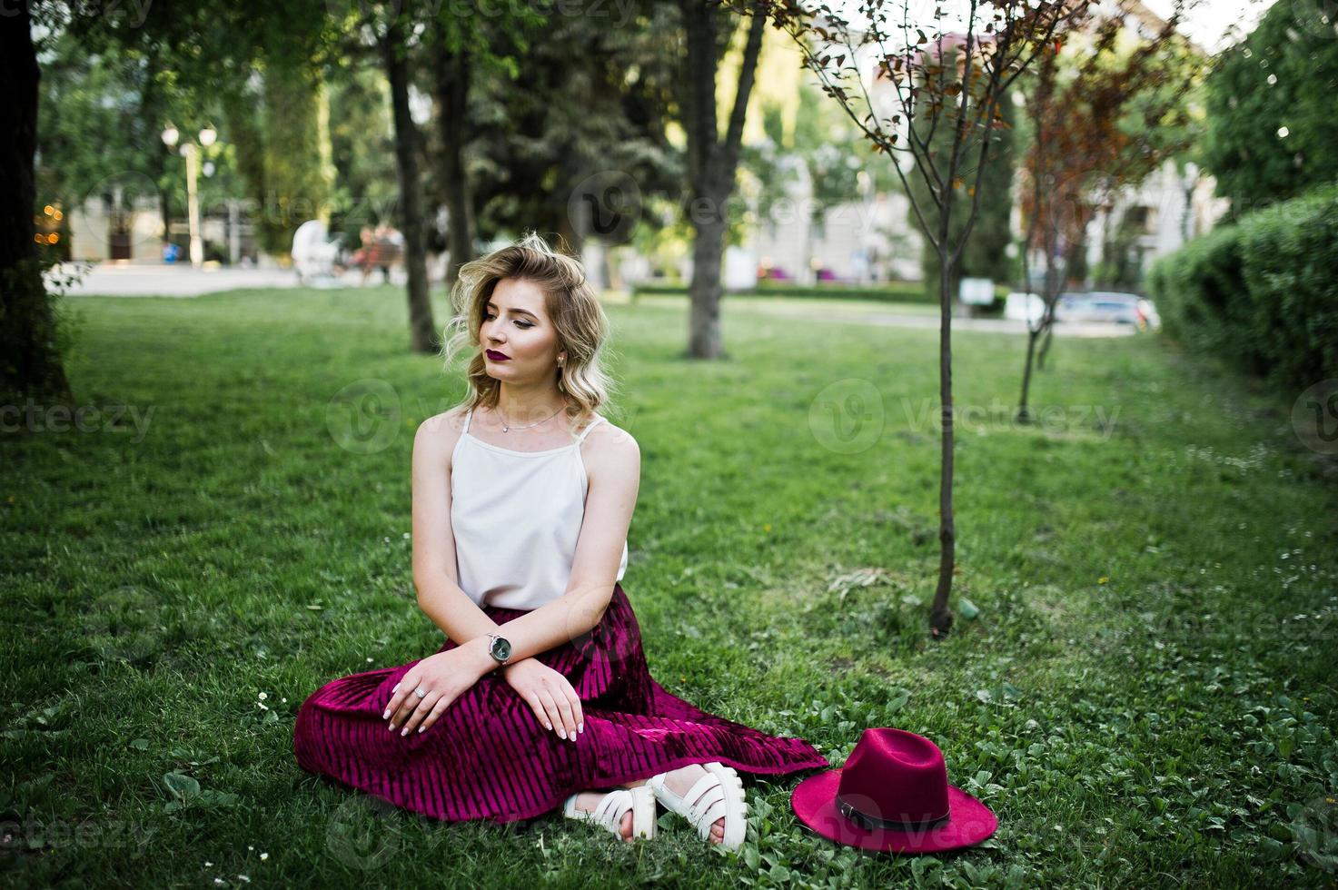 Fashionable and beautiful blonde model girl in stylish red velvet velour skirt, white blouse and hat, sitting on green grass at park. photo