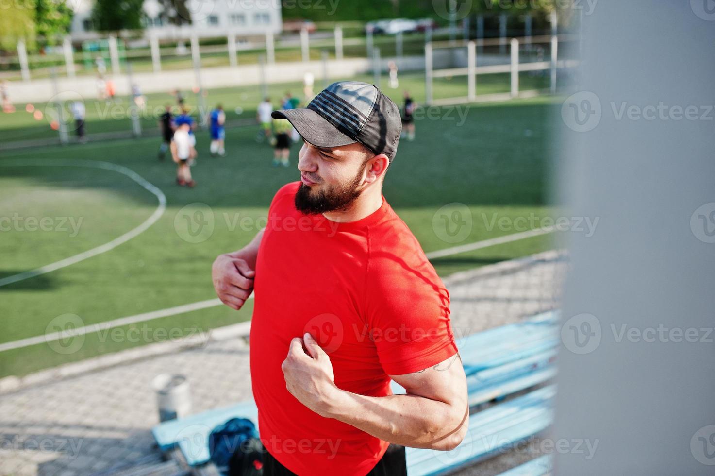 Young brutal bearded muscular man wear on red shirt, shorts and cap at stadium. photo