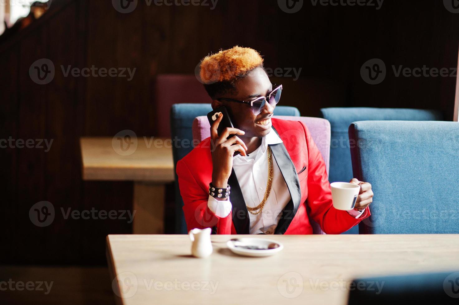 Fashion african american man model at red suit, with highlights hair and sunglasses sitting at cafe with coffee and speaking on mobile phone. photo
