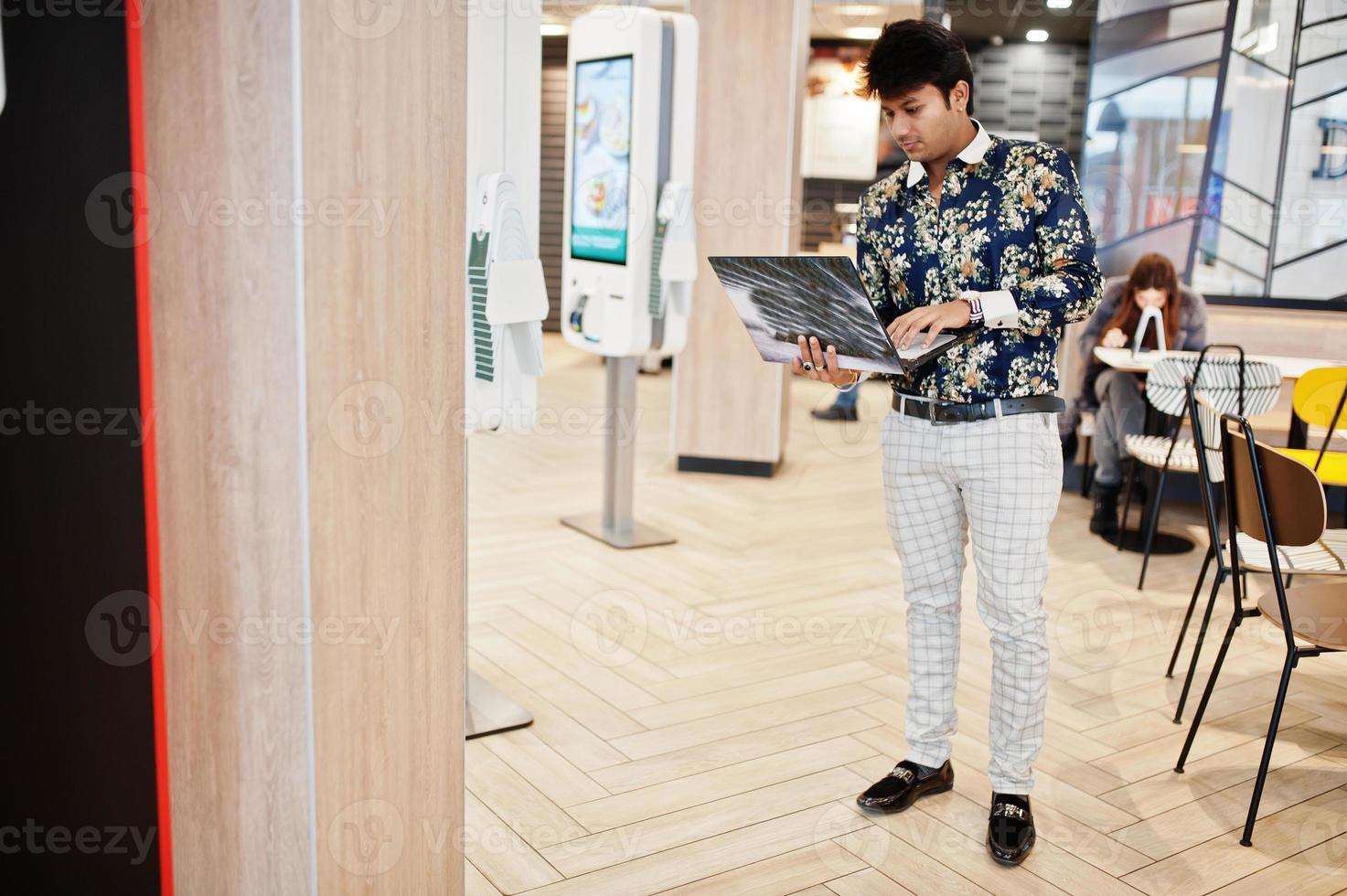 Young male indian freelancer sitting open laptop computer in fast food cafe, handsome asian man reading text message during work on net-book in comfortable coffee shop. photo