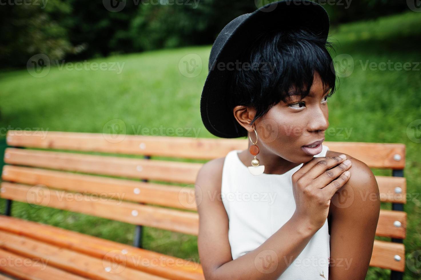 Increíble mujer modelo afroamericana con pantalones verdes y sombrero negro posada en un banco en el parque. foto