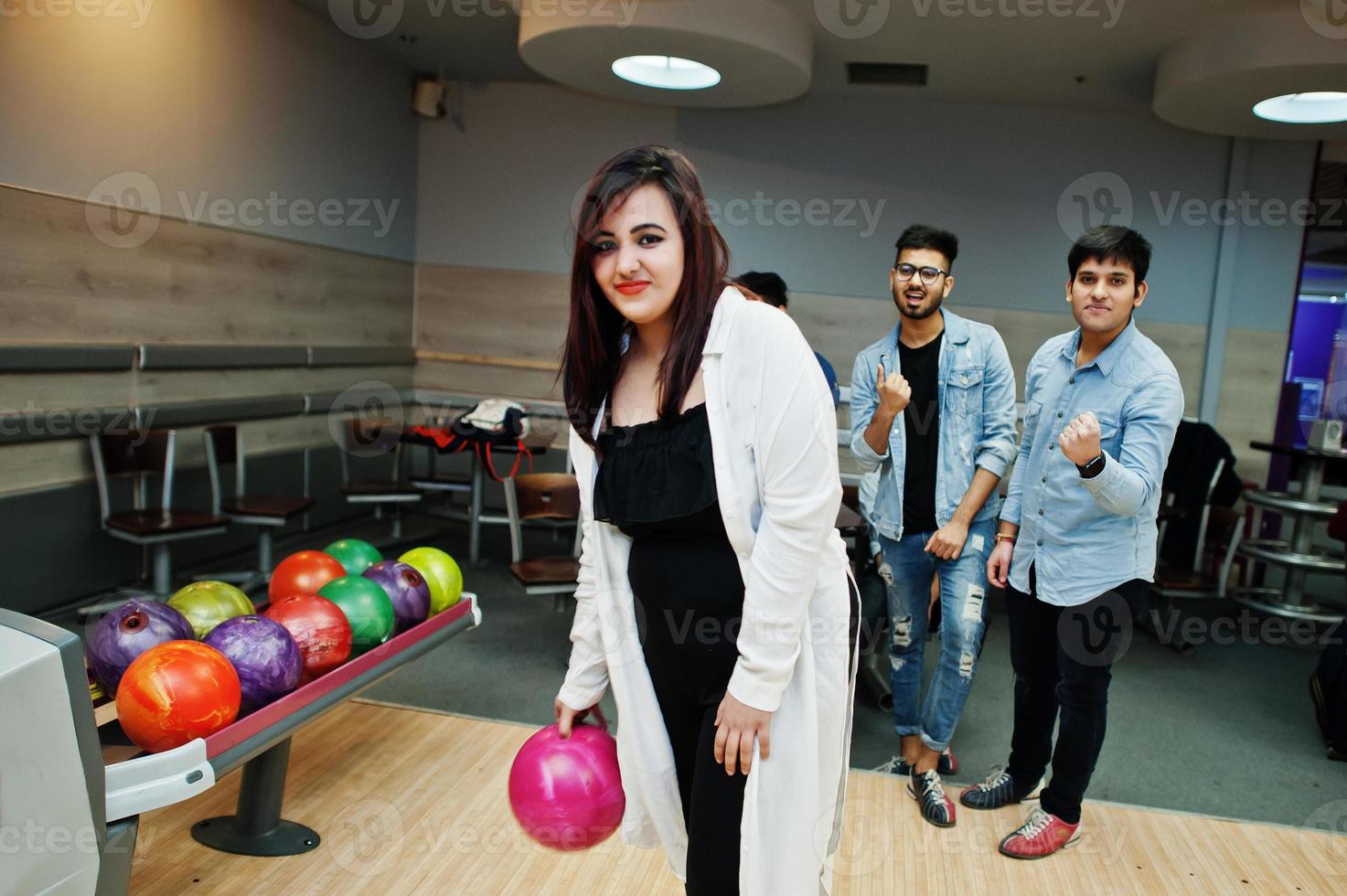 mujer del sur de asia parada en una bolera con pelota en las manos. la chica se está preparando para un lanzamiento. los amigos la apoyan en voz alta. foto