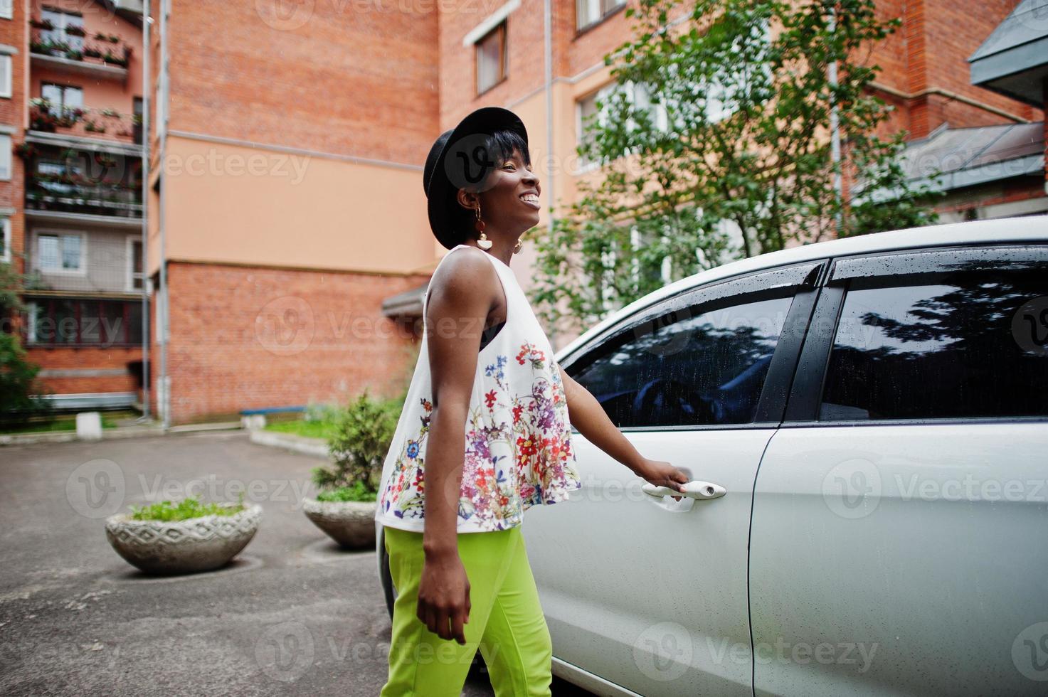 Amazing african american model woman in green pants and black hat posed near white door of car. photo