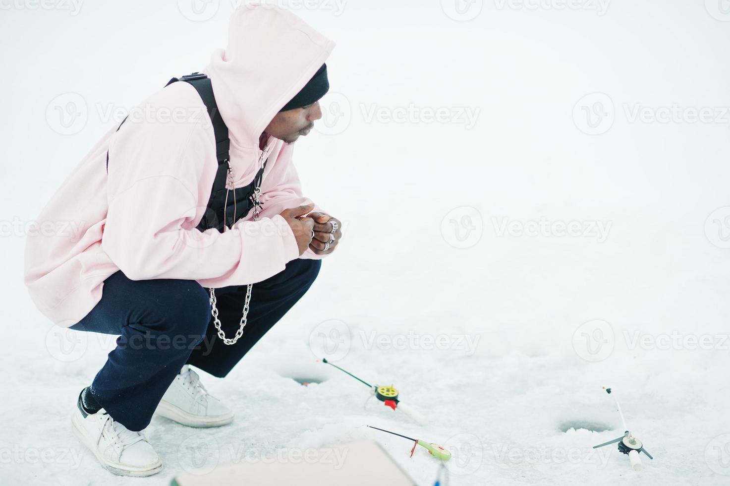 African american fisherman with fishing rod sitting on frozen sea. Winter fishing. photo