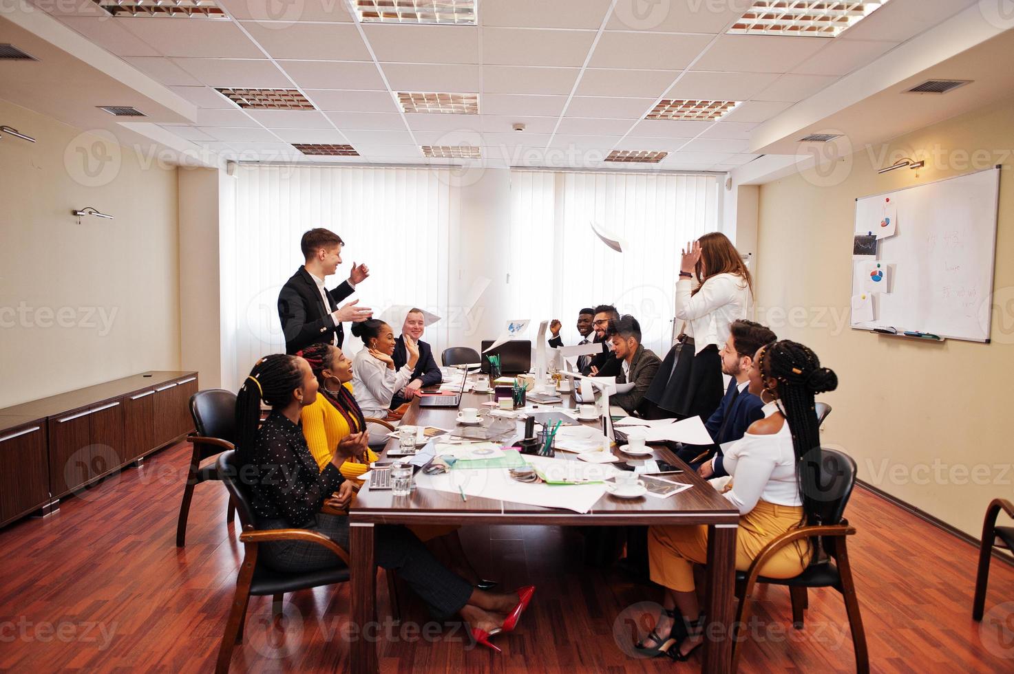 Multiracial business team meeting around boardroom table, two team leaders throw paper up. photo