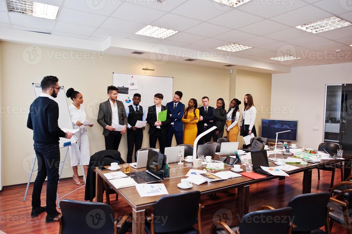 Mixed race business coach presenting report standing near whiteboard pointing on sales statistic shown on diagram and chart teach diverse company members gathered together in conference room. photo