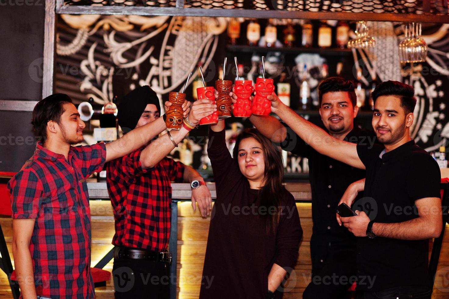 Group of indian friends having fun and rest at night club, drinking cocktails, clinking and cheers near bar counter. photo