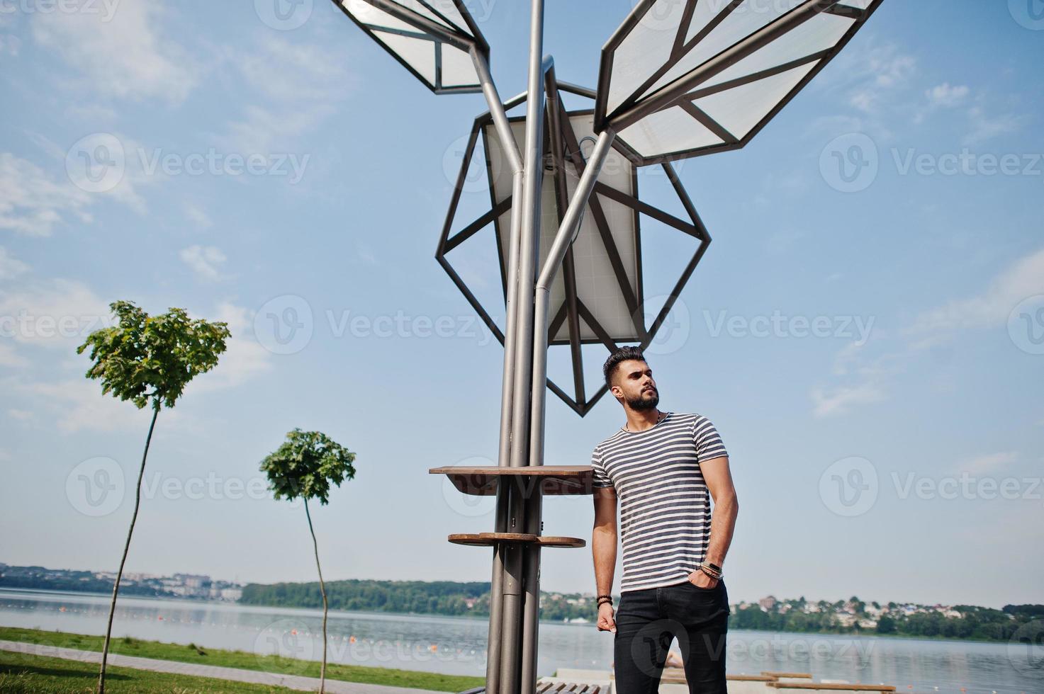 Handsome tall arabian beard man model at stripped shirt posed outdoor against solar panel. Fashionable arab guy. photo