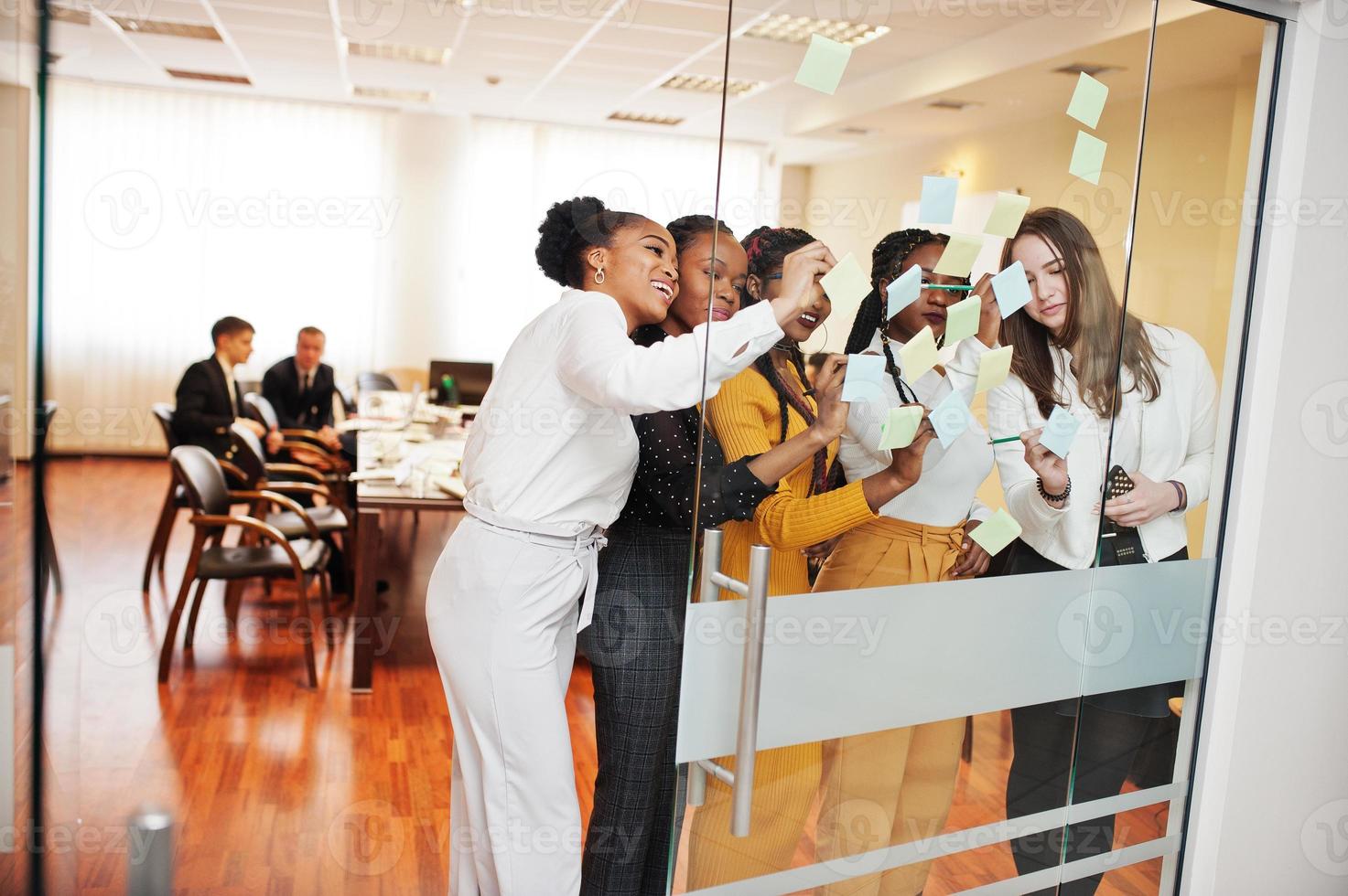 Business womans pointing on glass with colorful paper notes. Diverse group of female employees in formal wear using stickers. photo