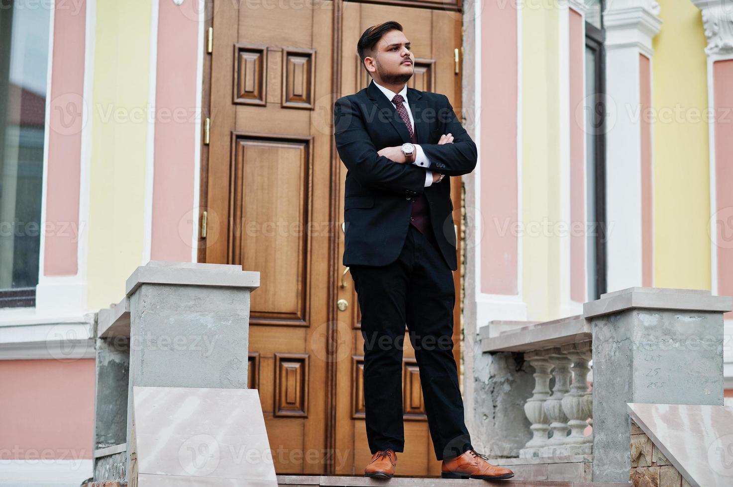 Stylish indian businessman in formal wear standing against door in business center with cross arms. photo