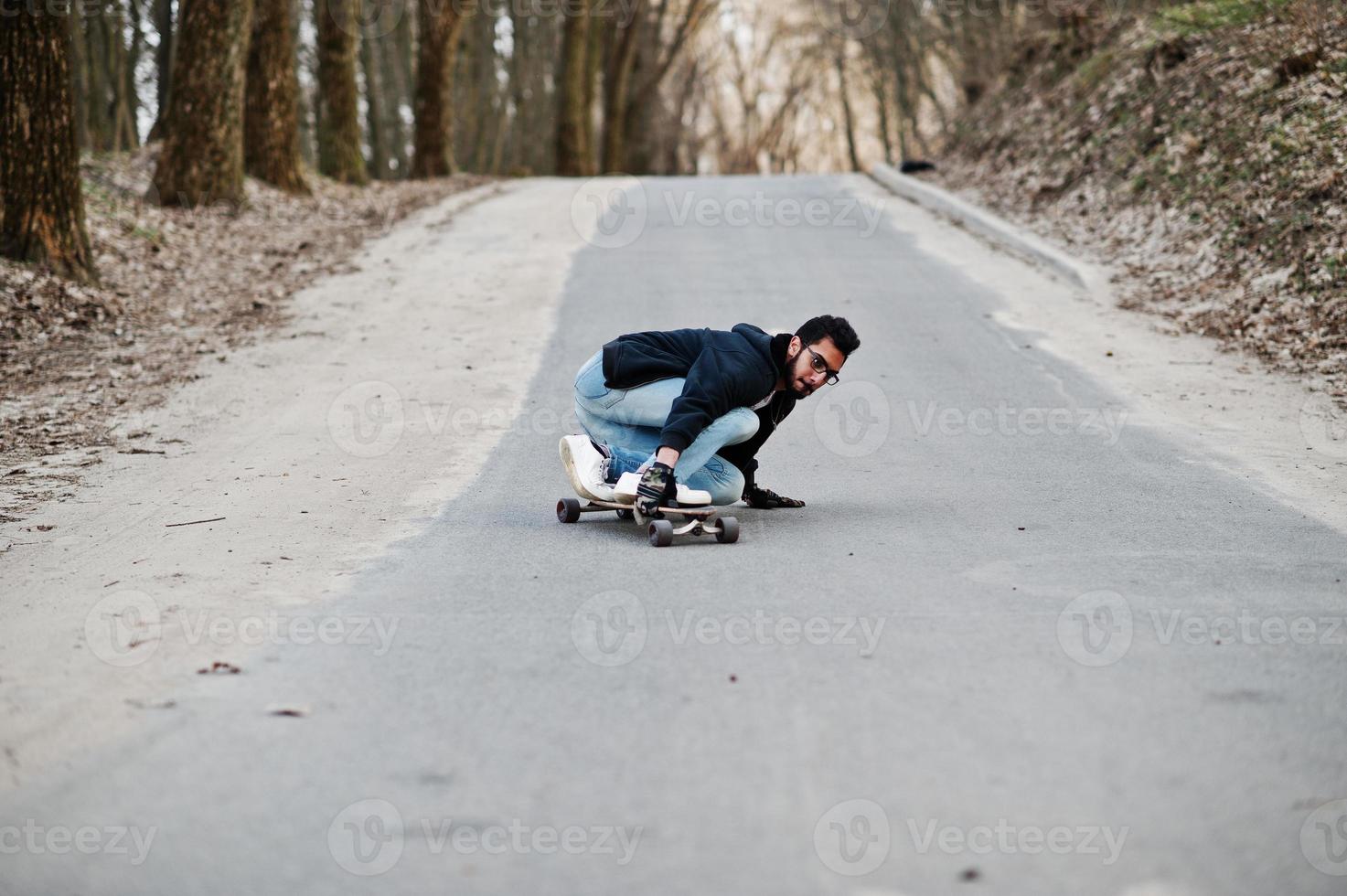 hombre árabe de estilo callejero en anteojos con longboard longboarding por el camino. foto